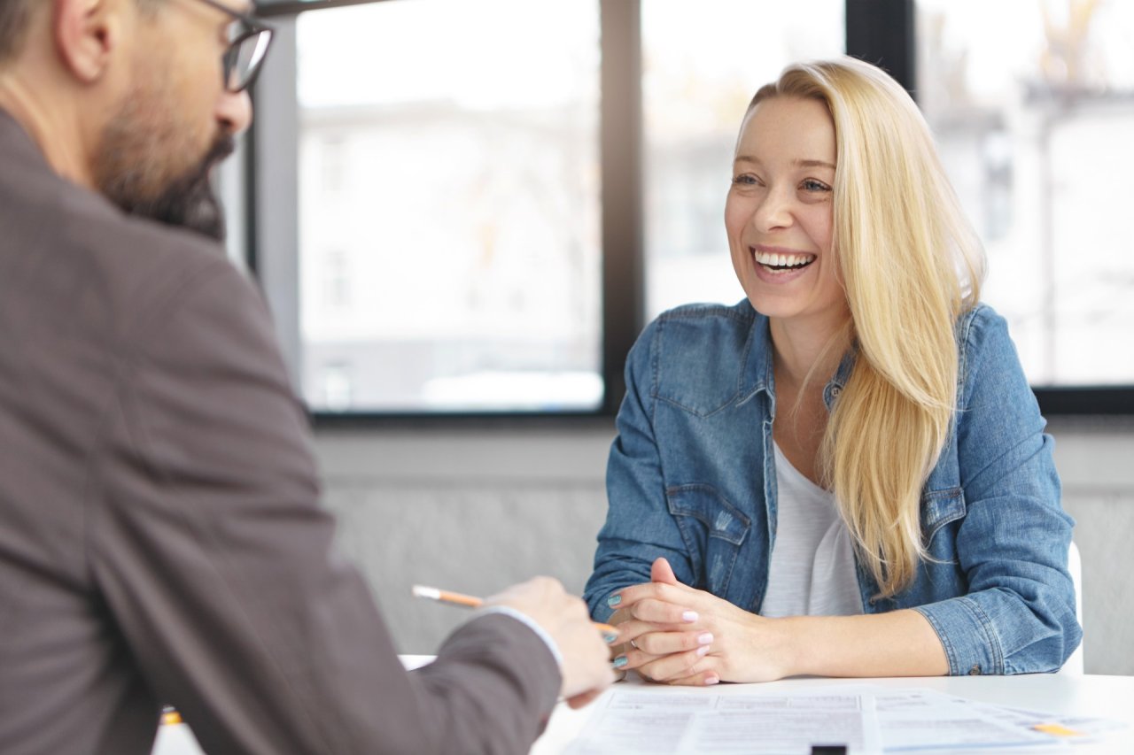 blonde woman and man with glasses, sat talking, looking over work notes, smiling.