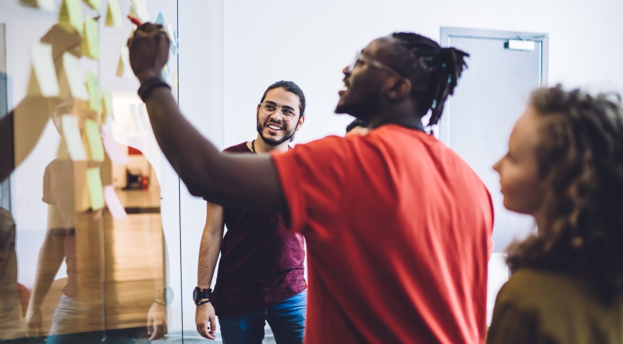 group of employees in casual dress, stood in front of a glass whiteboard with post in notes on.