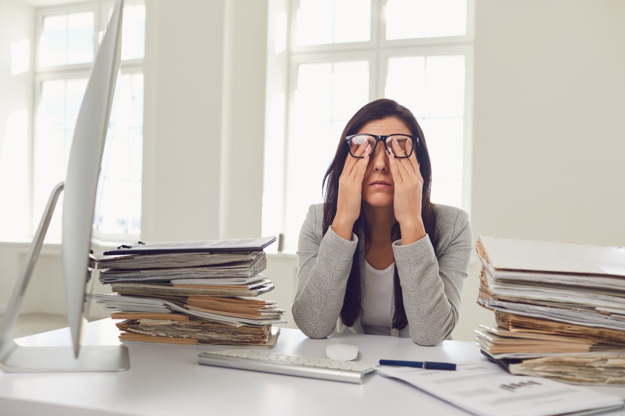 Woman sat at work desk, rubbing her eyes, surrounded by paperwork.